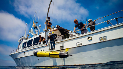 The Sentinel Redwing glider, a yellow submersible research robot is hoisted from a ship to bring it into the ocean.