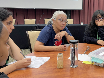 Several young people sitting at a round table with an older woman. They listen in as she speaks. 