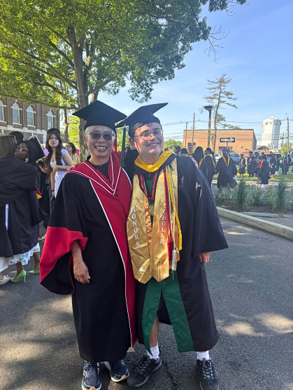 Two people, a professor and a student, posing in convocation regalia on a college campus. Behind them are other graduates and their loved ones. 