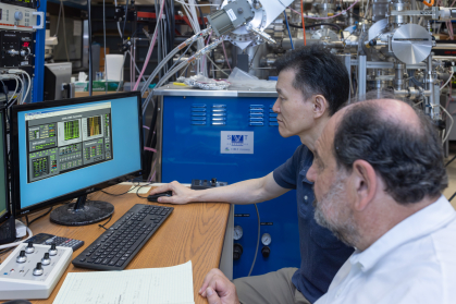 Two men seated observe images on the screen of a computer. In the background are lots of pipes and wires.