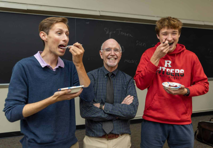 A professor stands in the middle of two students who are eating cranberries